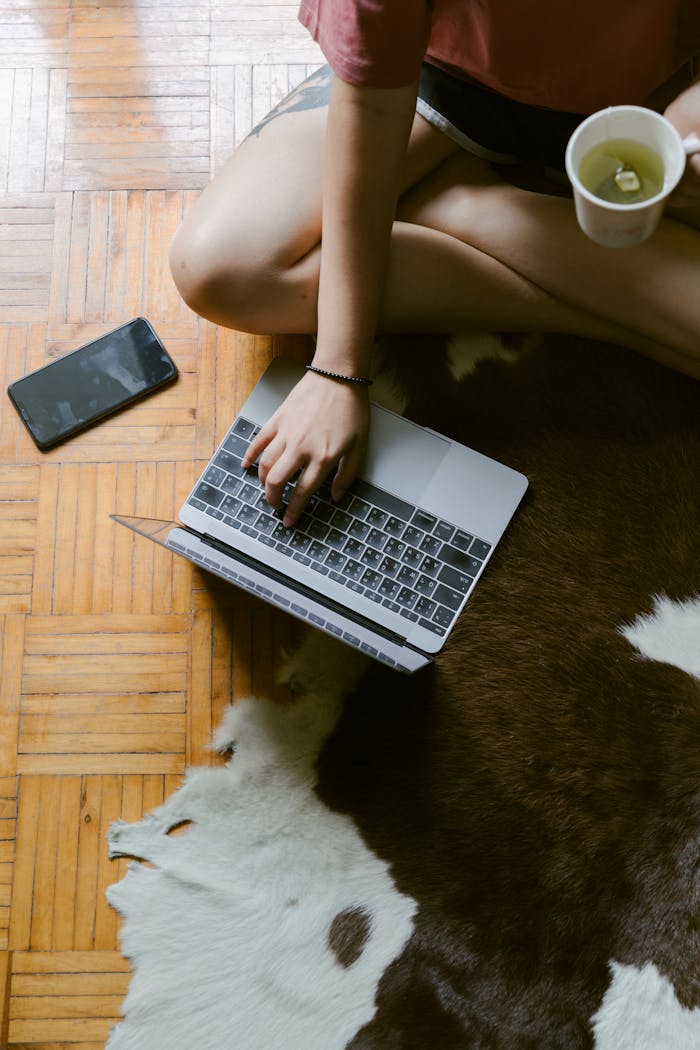Top view of faceless female using laptop while sitting on floor with cozy carpet with cup of  green tea